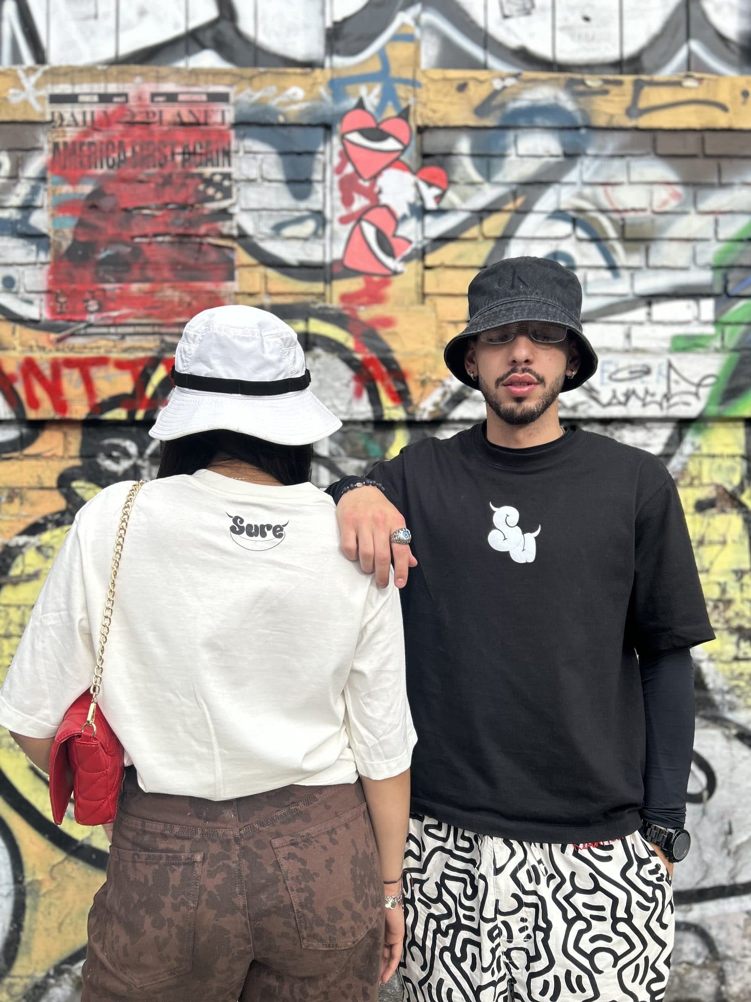 Pareja frente a un muro de grafiti con las camisetas Intrinsic blanca y negra
