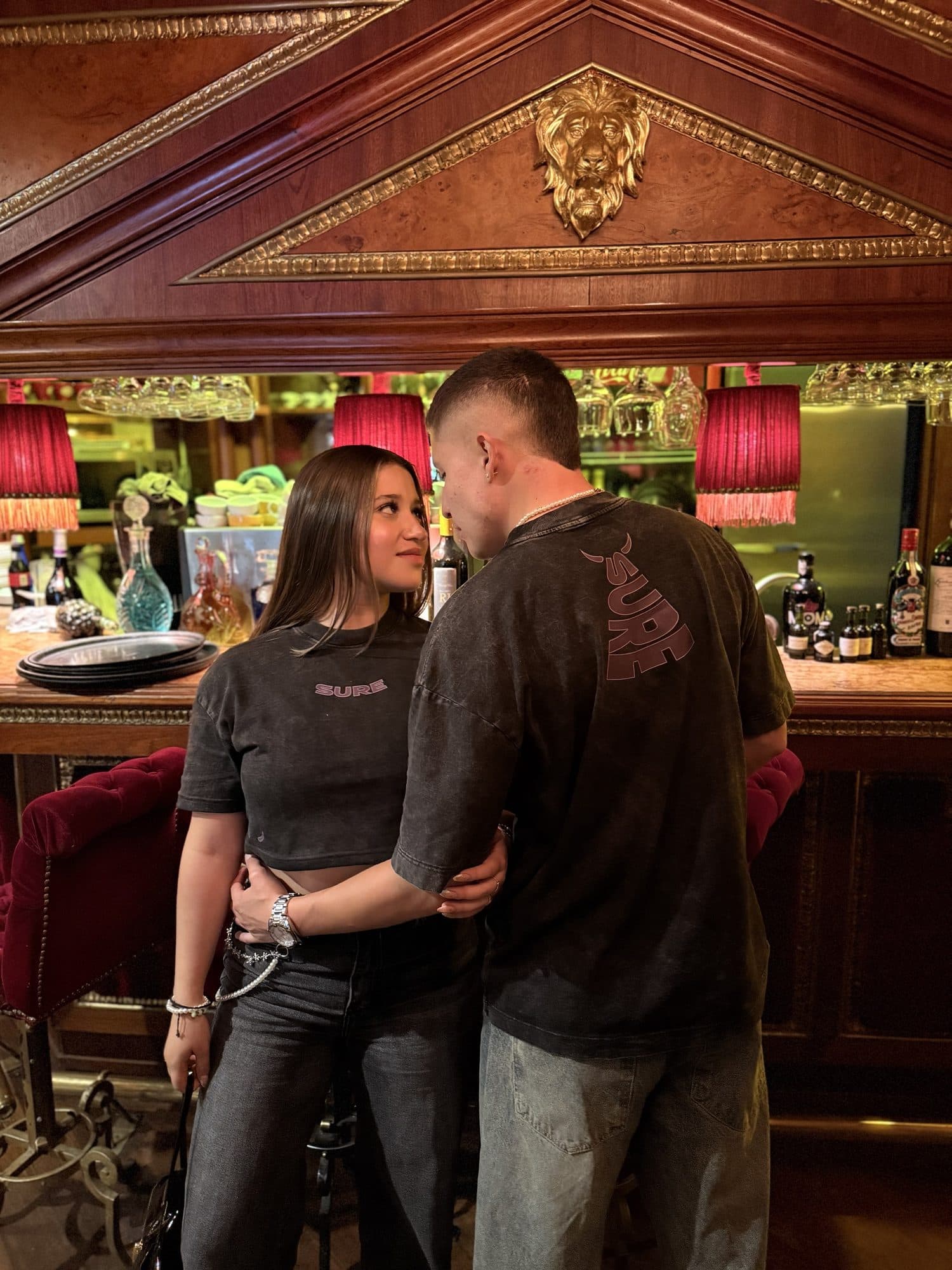 Pareja en un bar con la camiseta y el crop gris carbón; atrás, SURE con cuernos en morado