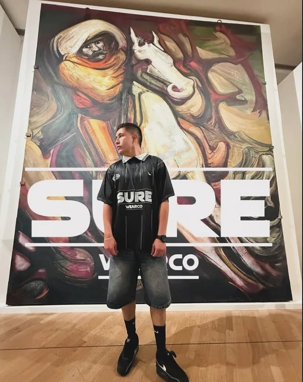 Modelo con la camiseta Sure26 frente a un mural con el logo SURE