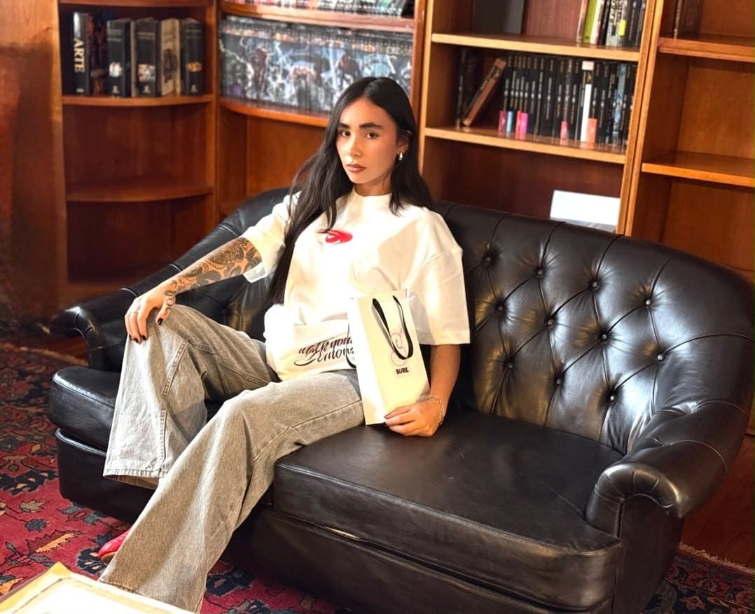 Modelo con la camiseta Disidencia blanca y una bolsa de SURE en las manos, sentada en un sofá de cuero